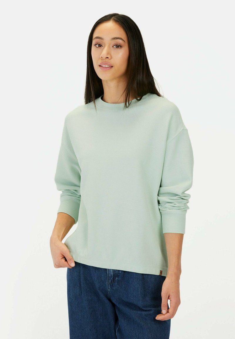 Mintgroene sweatshirt met textuur, ronde hals, losse pasvorm en lange mouwen, gecombineerd met donkere spijkerbroeken. Subtiele branding zichtbaar.