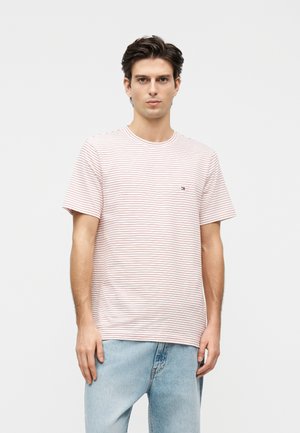 Young man standing, wearing a white t-shirt with thin red stripes and light blue jeans, against a plain light gray background.