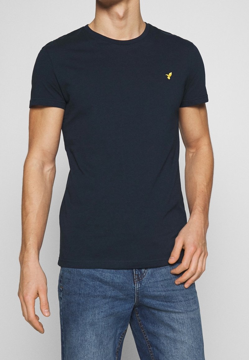 Homme portant un t-shirt bleu marine ajusté avec un petit logo jaune sur la poitrine et un jean bleu, debout devant un fond uni clair.