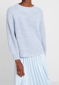 Hellblauer gestrickter Pullover mit rundem Halsausschnitt, gerippten Bündchen und lockerer Passform, kombiniert mit einem plissierten hellblauen Rock.