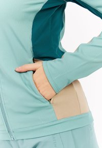 Ruka v vrecku svetlo modrej a béžovej zip-up bundy s kontrastným tmavým teal materiálom po bokoch, oblečená s rovnakými svetlo modrými nohavicami.