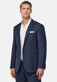 Blazer bleu marine en tissu texturé avec deux poches avant, fermeture à un bouton, revers crantés et pochette blanche.