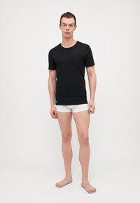 T-shirt nera a maniche corte con scollatura rotonda, realizzata in un materiale morbido, abbinata a pantaloni boxer bianchi e piedi nudi.