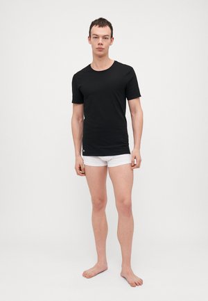 T-shirt noir à manches courtes avec un col rond, fabriqué à partir d'un matériau doux, associé à un short boxeur blanc et pieds nus.