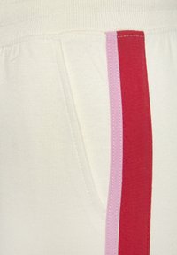 Weiße Jogginghosen mit einem seitlichen Streifenmuster in Rot, Rosa und Hellrose. Der Stoff hat ein weiches, strukturiertes Aussehen.