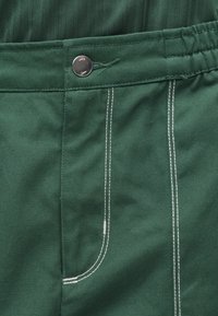 Tmavozelený pás na nohaviciach z látky so strieborným gombíkom a bielym dvojitým prešitím pozdĺž vrecka a švov.