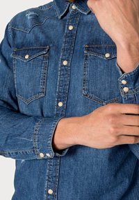 Man past dat een mouw van een blauw denim overhemd met drukknoopsluitingen en borstzakken.