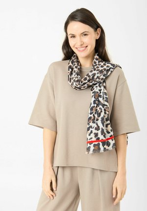 Vrouw draagt beige loszittende top en broek met een luipaardprint sjaal met een rode streep, glimlachend tegen een effen witte achtergrond.