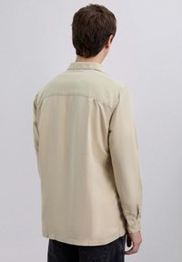 Camisa de manga larga color beige claro con cuello, textura suave, puños con botones y detalles en la parte trasera. Combinada con pantalones negros.