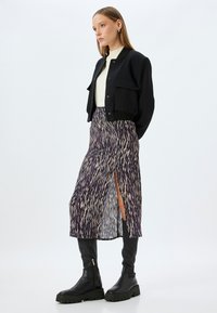 Veste noire courte avec fermeture à boutons, chemise crème, et jupe midi violette à motif avec une fente sur le côté, associée à des bottes noires jusqu'aux genoux.