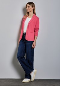 Różowa teksturowana marynarka z dwoma guzikami, zestawiona z białą bluzką i szerokimi, ciemnoniebieskimi dżinsami. Całość dopełniają białe trampki.