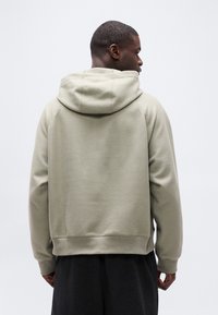 Άνδρας με πλάτη, φοράει ανοιχτόμπεζ φούτερ με κουκούλα και μαύρο παντελόνι, σε απλό λευκό φόντο.