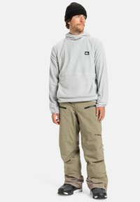 Pull en polaire gris clair avec un col haut et un petit logo, associé à un pantalon de neige beige avec des poches zippées et des accents noirs.