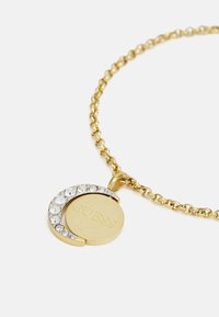 Collier en or avec un pendentif rond orné de cristaux sur un bord et un logo gravé. La chaîne est composée de maillons en forme d'ovale.