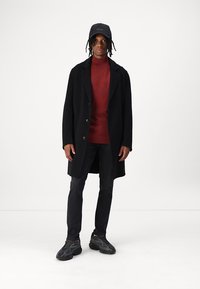 Sort frakke med store knapper, rød strikket turtleneck, mørke jeans og mørke sneakers. Kasket med "BOSS" logo fuldender outfittet.
