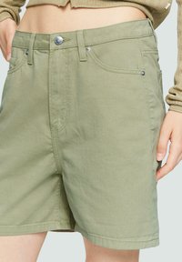 Shorts vert olive en coton, avec une fermeture à bouton sur le devant, des passants de ceinture, des poches latérales et une coupe droite avec des coutures visibles.
