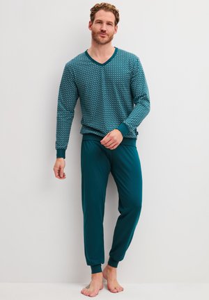 Man met krullend haar, gekleed in een teal gekleurd langermig shirt met patroon en bijpassende teal joggerbroek, staat blootsvoets tegen een effen muur.