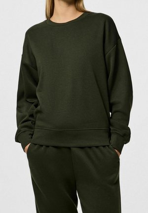 Ciemnozielona bluza z okrągłym dekoltem, opadającymi ramionami i ściągaczami. Wykonana z miękkiego, teksturowanego materiału o luźnym kroju.