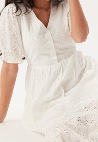 Biała sukienka z bufiastymi rękawami, z haftowanymi kwiatowymi wycięciami, dekoltem w kształcie litery V i detalami w postaci guzików, wykonana z lekkiego materiału.