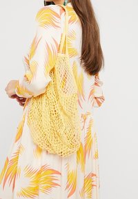 Geel, netvormige schoudertas met een zachte textuur en opengewerkt ontwerp, gedragen over de schouder, gecombineerd met een jurk met een patroon in pasteltinten.