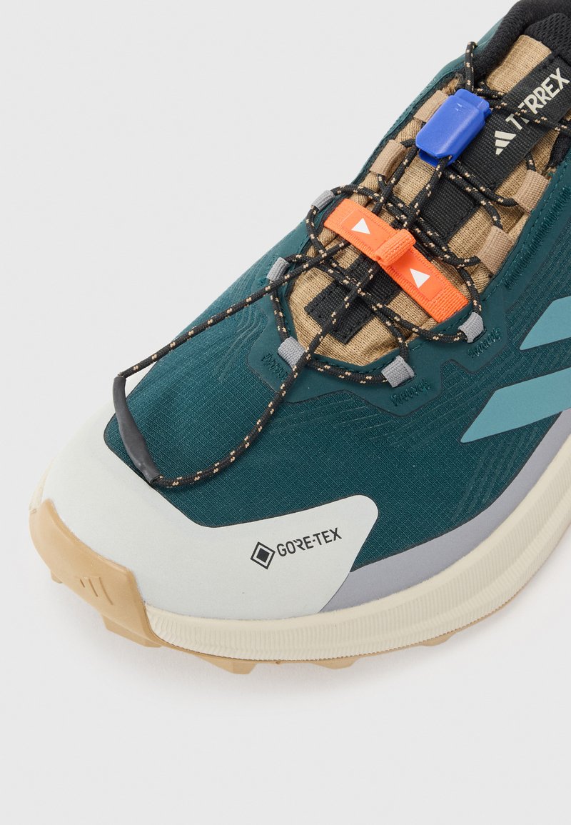 Zapatilla de trail running GORE-TEX en color teal y beige. Cuenta con tela texturizada, cordones elásticos, acentos en naranja y azul, y una suela acolchada.