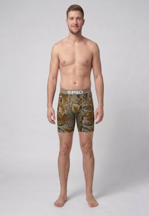 Junger Mann mit kurzem Haar steht barfuß vor einfachem Hintergrund und trägt PSD-Boxershorts im Camouflage-Muster, die Arme entspannt an den Seiten.