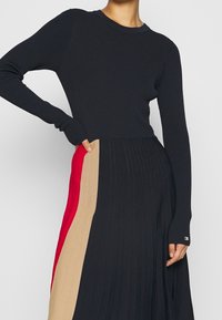 Robe en tricot à manches longues, avec un haut ajusté bleu marine foncé et une jupe plissée côtelée en coloris bloc noir, rouge et beige.