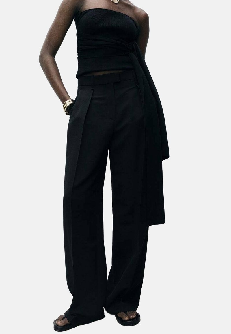 Femme portant un haut noir bustier avec une longue ceinture latérale et un pantalon noir à jambes larges, avec des sandales noires et des bracelets dorés.