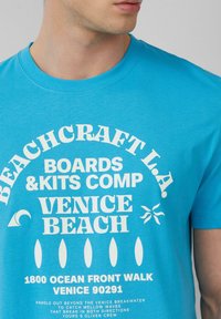Heldere blauwe katoenen T-shirt met witte bedrukte tekst, ronde halslijn en korte mouwen. Het ontwerp bevat strandthema-graphics en locatiegegevens.