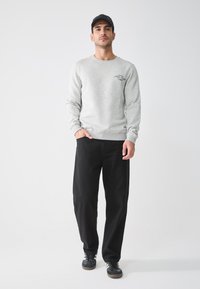 Sudadera gris claro con puños acanalados, que presenta un pequeño logo negro. Combinada con pantalones negros y zapatillas negras con rayas blancas.