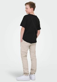 Czarny t-shirt z krótkim rękawem o zaokrąglonym dole, w połączeniu z beżowymi spodniami i białymi trampkami. Strój ma luźny krój i prosty design.