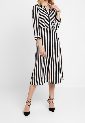 Kobieta ubrana w czarno-białą midi sukienkę na guziki z pionowymi paskami, z rękawami do połowy łokcia i czarne sznurowane szpilki.