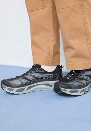 Czarne buty do biegania z siateczkowym i syntetycznym wierzchem, z szarymi akcentami i solidną podeszwą zapewniającą przyczepność. Noszone z beżowymi spodniami i białymi skarpetkami.