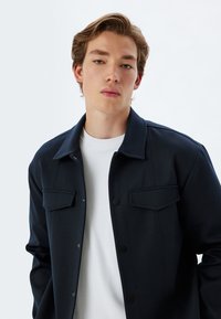 Mørkeblå knap-op jakke med to frontlommer, båret over en hvid crew neck skjorte, der har en glat tekstur og klassisk kravedesign.