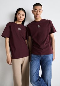 Burgundské trička s krátkým rukávem a bílým logem Lacoste, které nosí ženský a mužský model, v kombinaci s béžovými a modrými kalhotami.