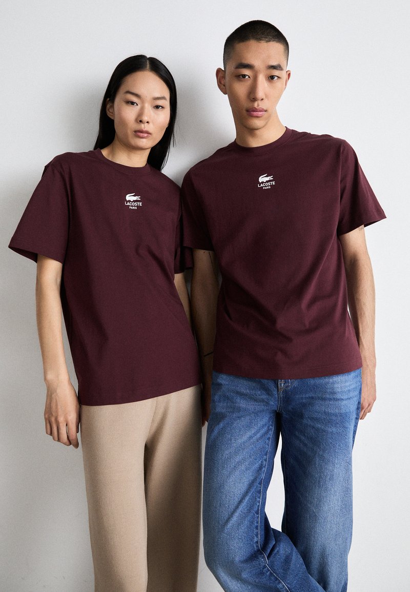 T-shirts bordeaux à manches courtes avec logo Lacoste blanc, portés par un modèle féminin et un modèle masculin, associés à des pantalons beiges et bleus, respectivement.