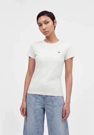 Femme aux cheveux courts et foncés portant un t-shirt blanc simple et ajusté ainsi qu'un jean large bleu clair, debout devant un fond blanc.