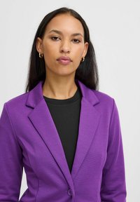 Femme aux longs cheveux bruns, portant de petites boucles d'oreilles en or, un blazer violet et un haut noir, regardant avec confiance devant elle contre un fond uni.