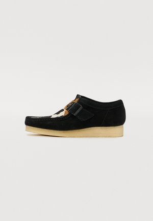Scarpa in suede nera con un design a stampa animale a contrasto nella parte anteriore. Presenta una fibbia regolabile e una suola in gomma di colore chiaro.