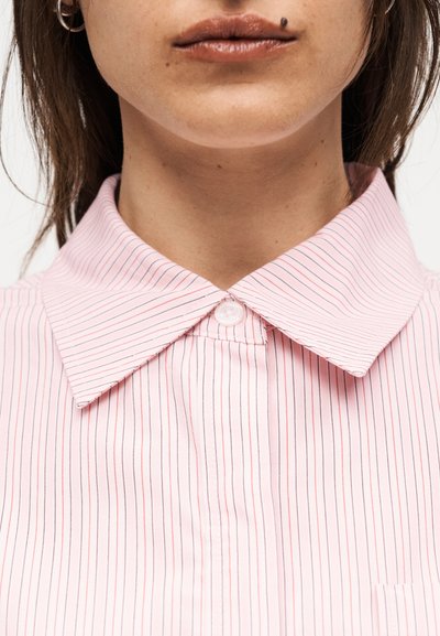 Γυναίκα που φορά ροζ και λευκό ριγέ πουκάμισο με γιακά, με το επάνω κουμπί κλειστό, εμφανές piercing στα χείλη και απλά σκουλαρίκια κρίκους.