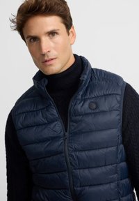 Hombre vestido con un chaleco sin mangas acolchado en color marino sobre un suéter de cuello alto de punto trenzado negro, mirando ligeramente hacia un lado.