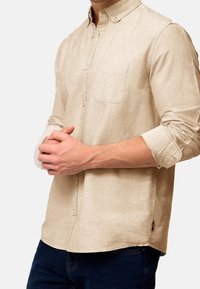 Camisa beige de botones hecha de tela texturizada, con un único bolsillo en el pecho y mangas enrollables, combinada con unos vaqueros oscuros.