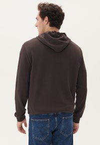 Hanorac maro cu şnur, având mâneci lungi și o croială lejeră. Asociat cu blugi din denim albastri, prezentând o vedere din spate.