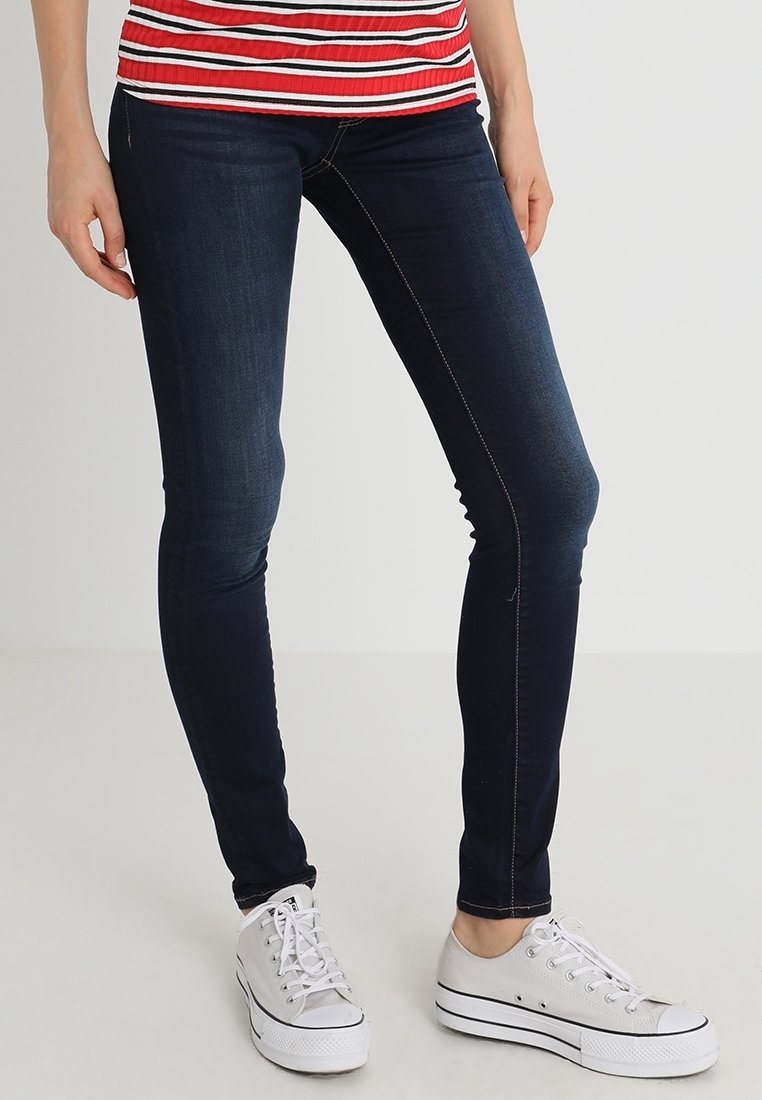 Vaqueros oscuros ajustados de color azul con una textura suave y costuras en contraste, combinados con zapatillas blancas de lona. Top de rayas rojas y blancas visible.