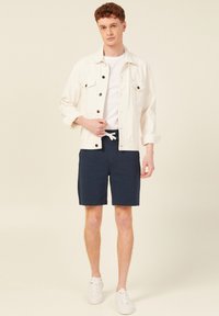 Biała jeansowa kurtka na białym T-shircie, zestawiona z granatowymi szortami i białymi sneakersami. Stylizacja charakteryzuje się swobodnym, luźnym krojem.