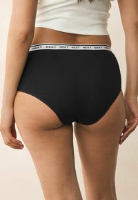 Black cotton brief with a white elastic waistband featuring the brand name "NEXT." High-rise design with smooth texture and full coverage.
