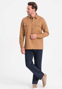 Bruine overhemd met knopen en twee borstzakken, gedragen met donkerblauwe spijkerbroek en beige sneakers. Glad materiaal, comfortabele pasvorm, casual ontwerp.