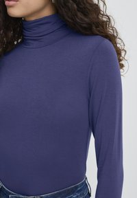 Donna dai capelli ricci che indossa un maglione attillato a maniche lunghe di colore viola e jeans blu, su uno sfondo bianco uniforme.