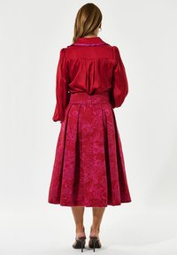 Rød satinbluse med bløde ærmer og en lilla krave; flydende rød nederdel med blomsterdesign, plisseret og med en tætsiddende talje.