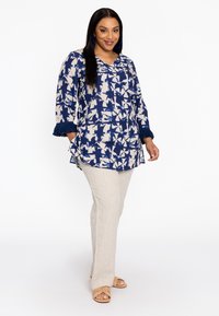 Navy en crèmekleurig patroon top met 3/4 mouwen en franjes, gecombineerd met lichtbeige broeken en tan sandalen.